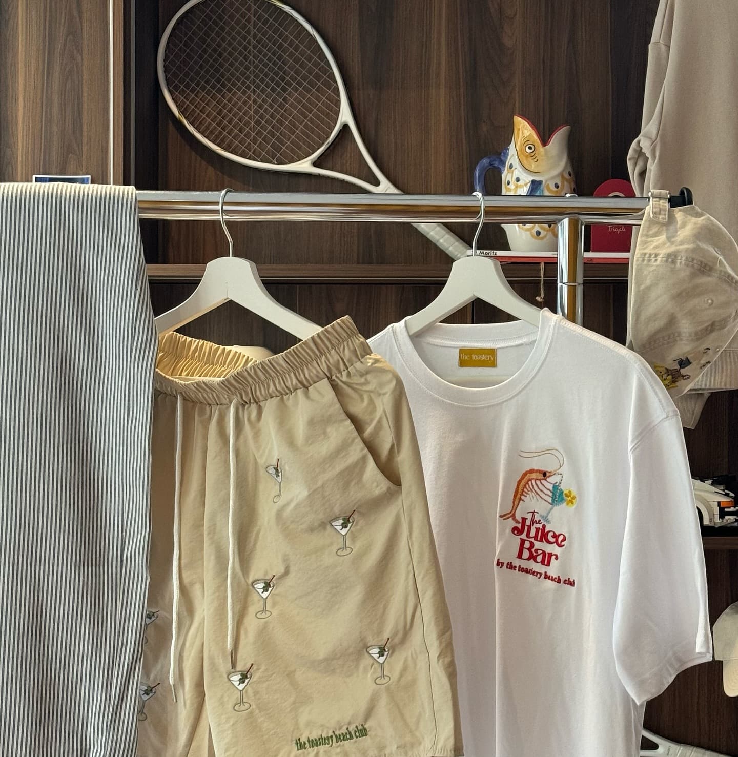 A chrome rail in front of walnut shelving: martini-embroidered shorts marked the toastery beach club, a white Juice Bar tee with a shrimp holding a cocktail, a cream cap and ticking-stripe trousers, with a tennis racquet and a painted fish jug on the shelf above.