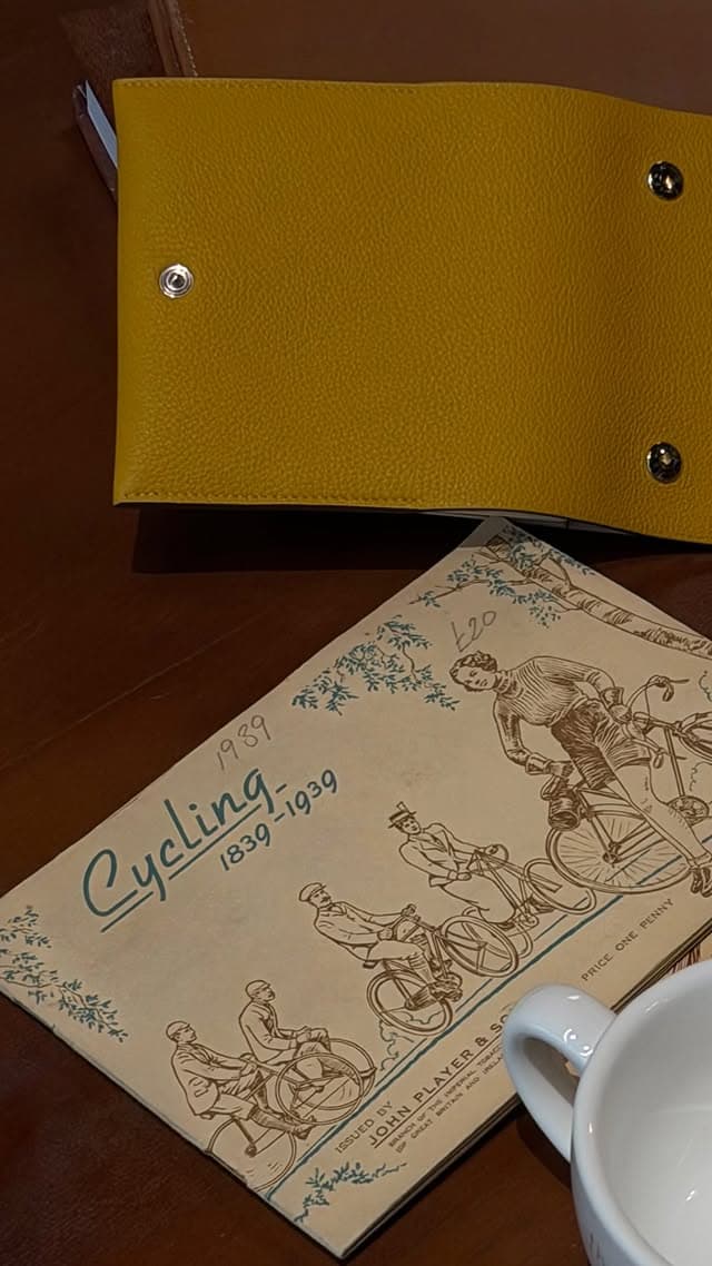 A mustard leather snap-close menu folder on a wooden table beside a 1939 John Player and Sons cigarette-card album titled Cycling 1839–1939, next to a white cup.