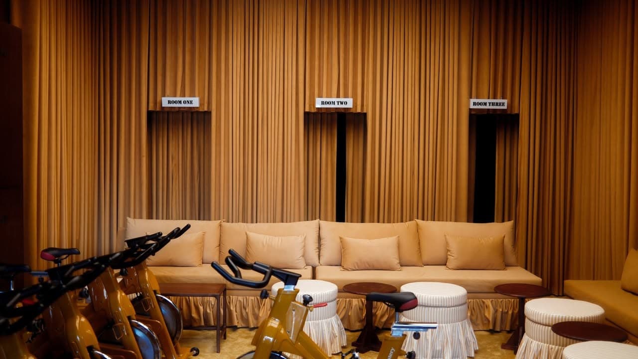The cycling studio: floor-to-ceiling ochre curtains with three recessed alcoves labelled ROOM ONE, ROOM TWO and ROOM THREE, a long mustard banquette with cushions, spin bikes in the foreground and striped skirted stools alongside.