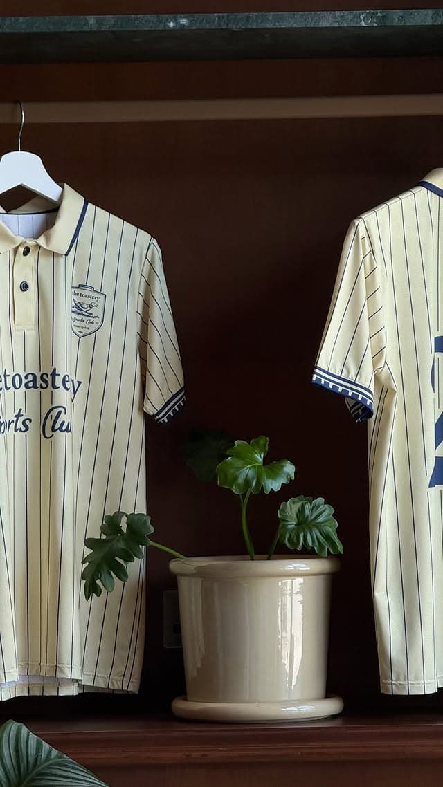 Two cream polo shirts with fine navy pinstripes hanging on a walnut shelf, one showing an embroidered shield crest on the chest, the other showing script lettering across the back, with a potted plant between them.