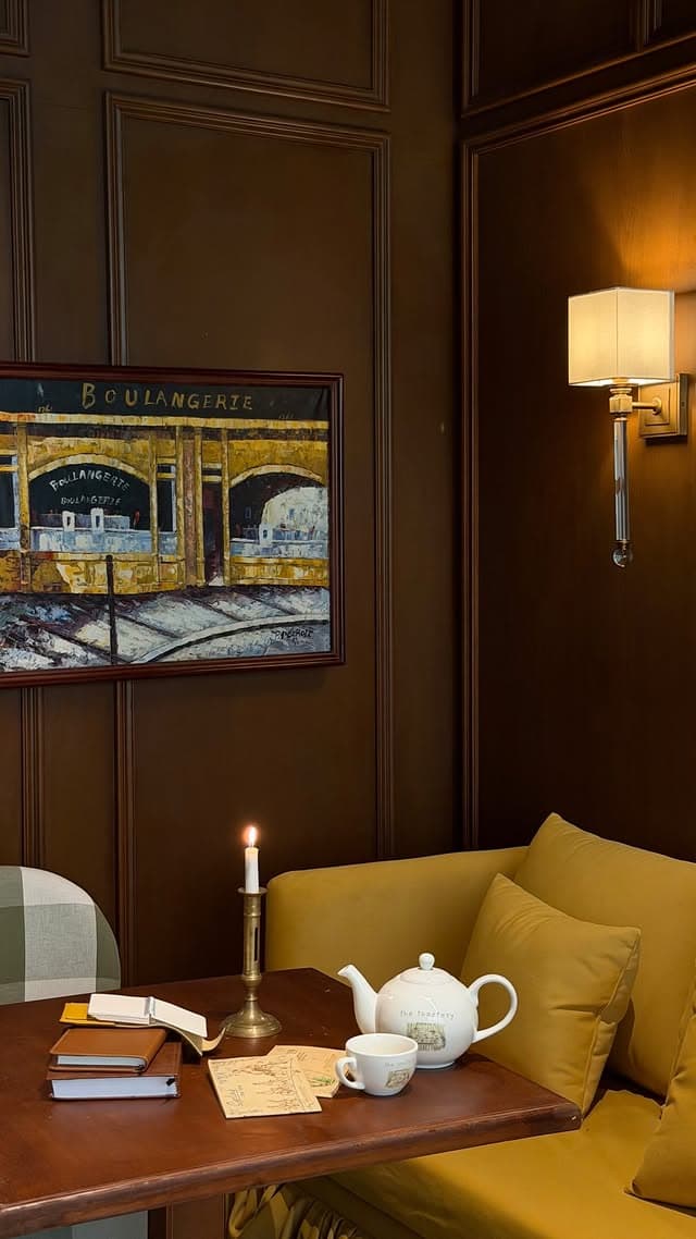 A corner banquette in mustard velvet beneath a painting of a boulangerie shopfront, with a lit candle, notebooks, and a branded white teapot on the table.