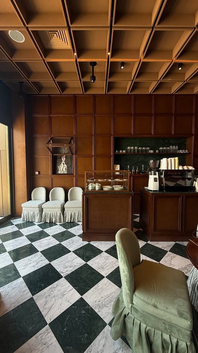 The café floor in green-and-white marble checkerboard, leading to a walnut-panelled bar with a pastry vitrine, espresso machine, and striped slipcovered chairs.