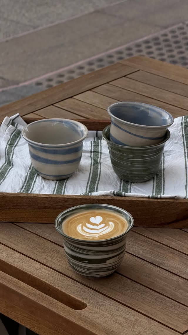 Three marbled ceramic funjans on a wooden tray with green-striped linen, and a fourth on the table holding a latte with rosetta art.