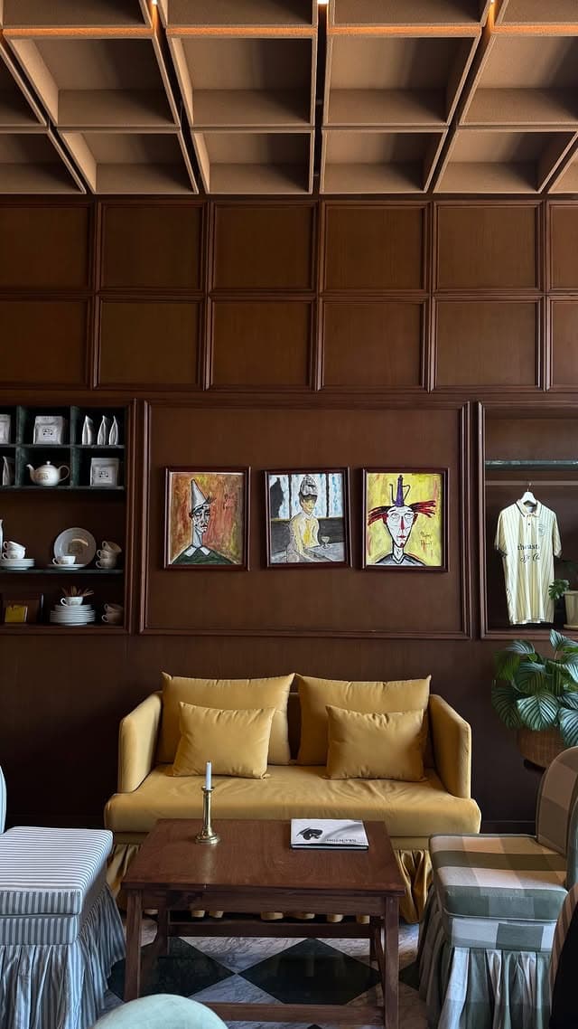 A mustard sofa below three framed portrait paintings, with shelves of white ceramics to the left and a pinstriped polo displayed in a lit vitrine to the right.