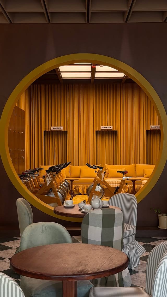 A café table set with a teapot, seen in front of an enormous gold-rimmed circular opening that frames the cycling studio beyond: ochre curtains, a row of spin bikes, and a mustard banquette.